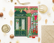 Lade das Bild in den Galerie-Viewer, Weihnachtskarte in einem Flatlay mit passendem Kraftpapier Umschlag und Weihnachtsdekoration. Die Grußkarte zeigt einen üppig weihnachtlich dekorierten Weihnachtsladen mit englischer Aufschrift merry and bright in festlichen Farben
