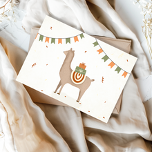 Lade das Bild in den Galerie-Viewer, Eine altweiße Karte zum Geburtstag mit einem braunen Alpaka, das Geschenke trägt und einen bunten Sattel trägt. Das Lama ist von Konfetti und einer Girlande aus orangefarbenen und grünen Flaggen umgeben. Die Karte liegt auf einem braunen Umschlag aus Kraftpapier.
